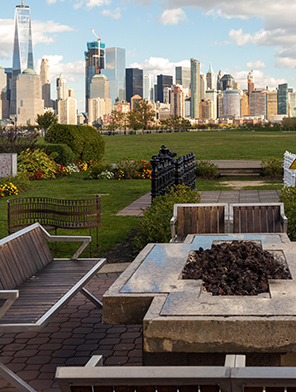 Professionally landscaped park with flowering plants and seats around a fire pit, NYC in background.